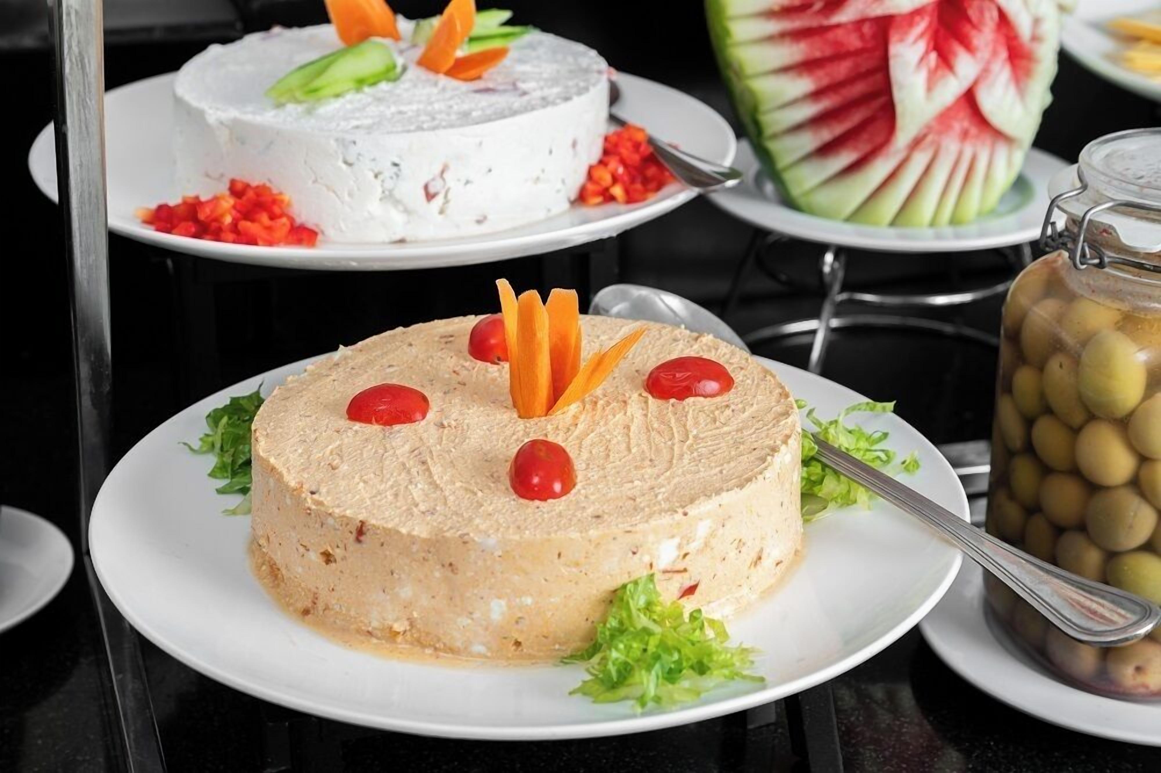 Een ronde tafel met een wit bord waarop een ronde cake ligt, versierd met wortel en kersen, een wit bord met een witte cake, en een watermeloen die is uitgehouwen tot een bloem.