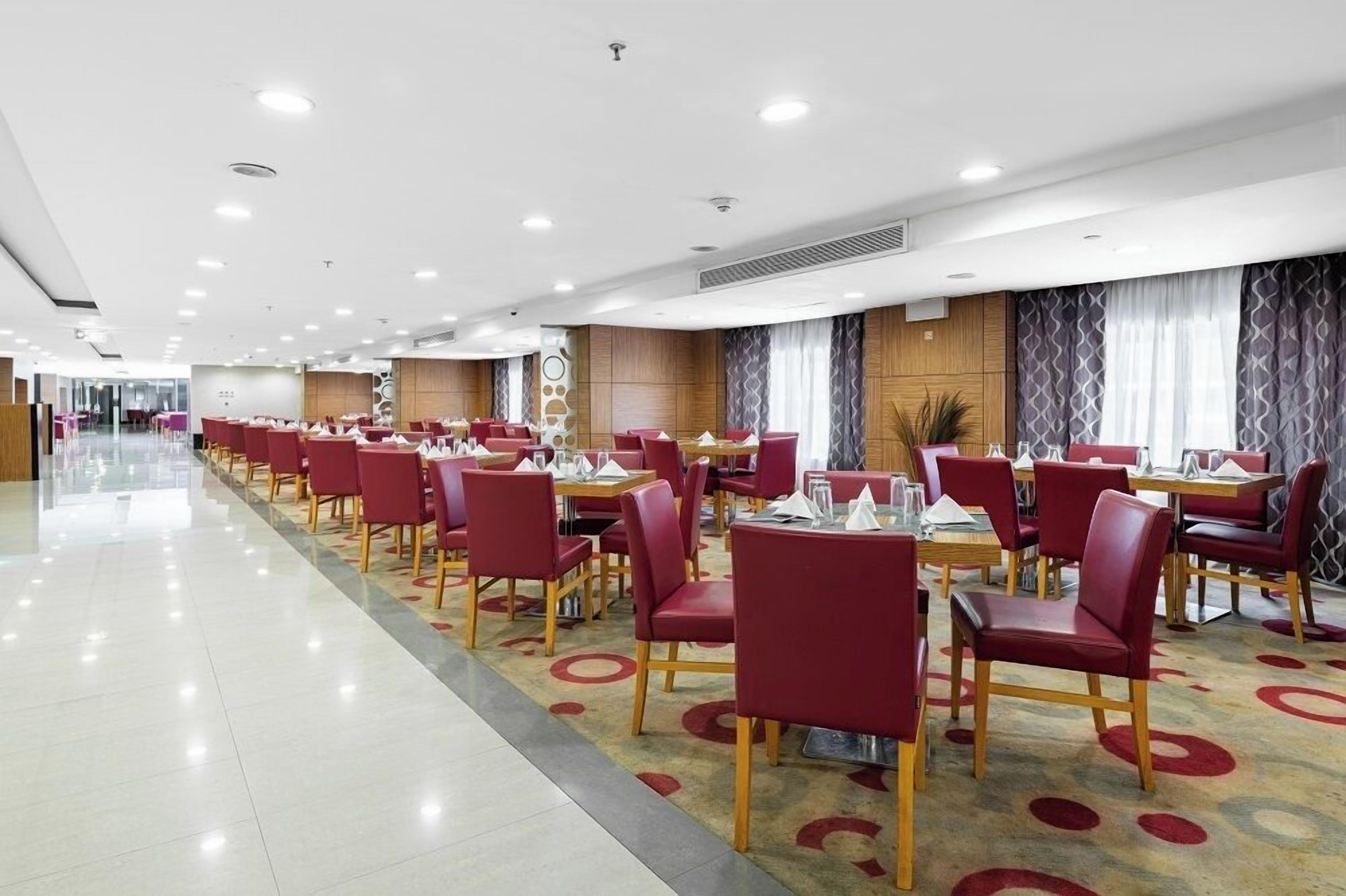 Un vaste espace de restauration d’hôtel, avec des chaises et des tables rouges, un sol ciré et un design moderne au plafond.