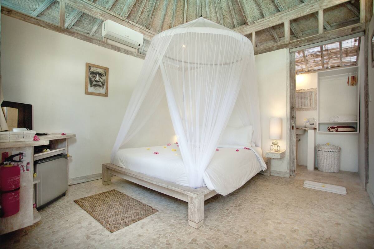 A bedroom with a canopy bed, a small desk with a chair, a bedside table with a lamp, and a wicker basket.