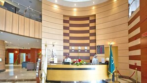 A hotel reception area with a curved wooden reception desk, a digital clock, and a flag. - Hotel Prestige (Makkah)