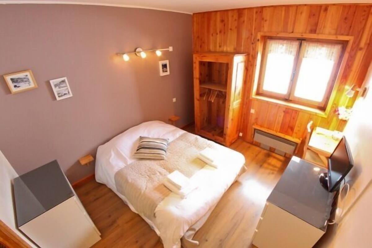 A bedroom with a bed, bedside tables, a wooden wardrobe, a window with curtains, and wall-mounted lights.