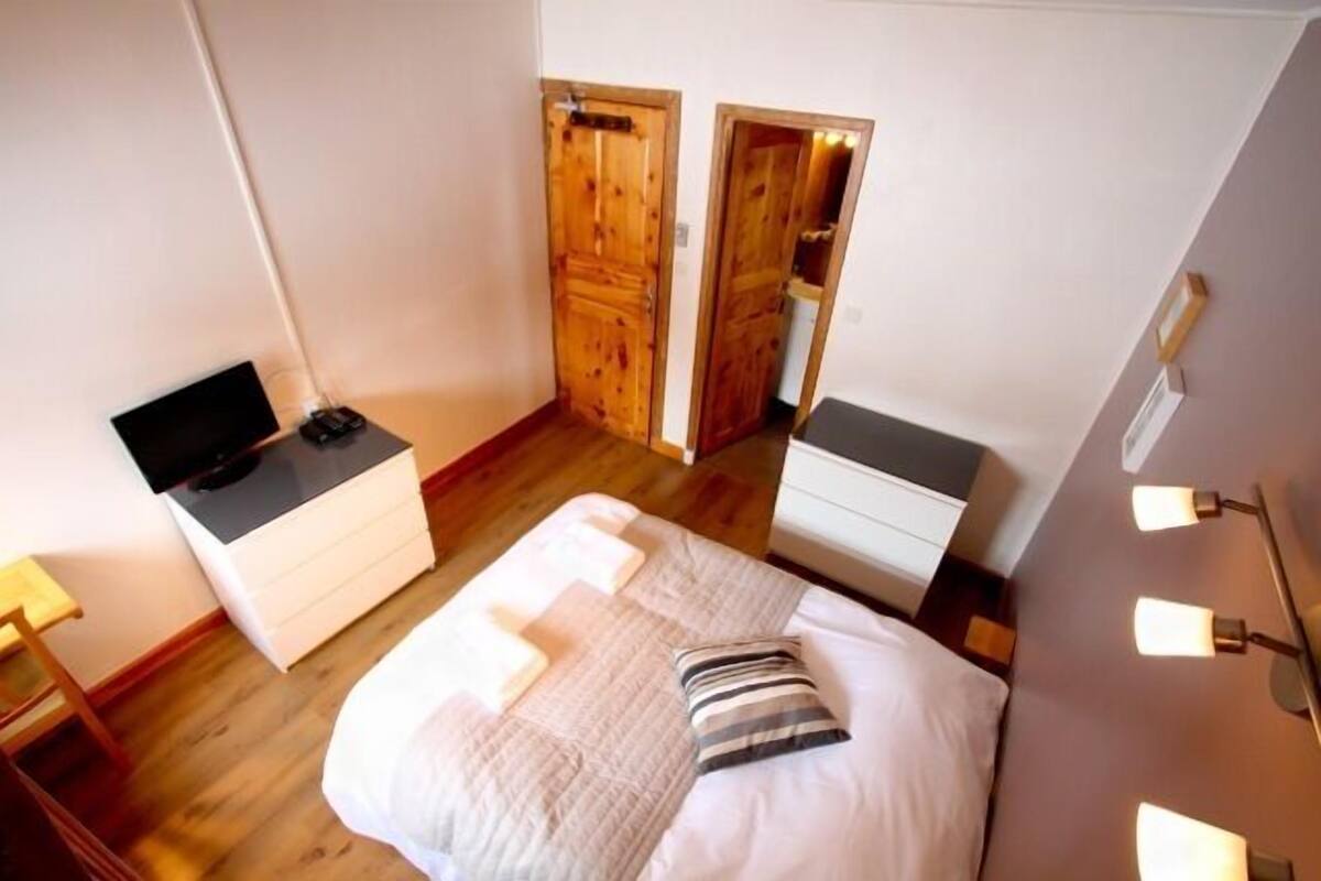 A bedroom with a bed, a desk, a chair, and a TV. There are wooden doors and a light fixture on the wall.