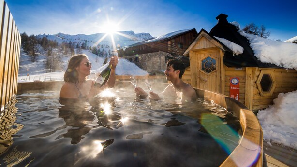 Un sauna en bois, dont le toit est recouvert de neige, entouré d’un paysage enneigé et de montagnes.