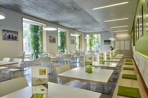 Ein moderner Hotel-Speisesaal mit weißen Tischen und Stühlen, großen Fenstern mit Grünpflanzen im Außenbereich und einem klaren, zeitgemäßen Design.