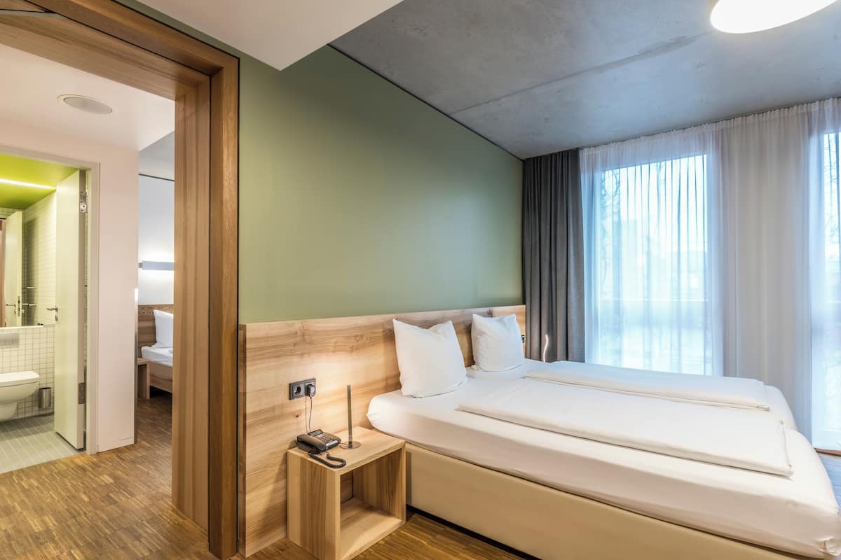 A modern hotel room with a wooden headboard, a bedside table with a phone, and a large window with curtains.