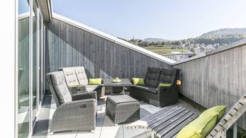 A rooftop patio with wicker furniture, a small table with a bowl of fruit, and a view of a distant town and hills.