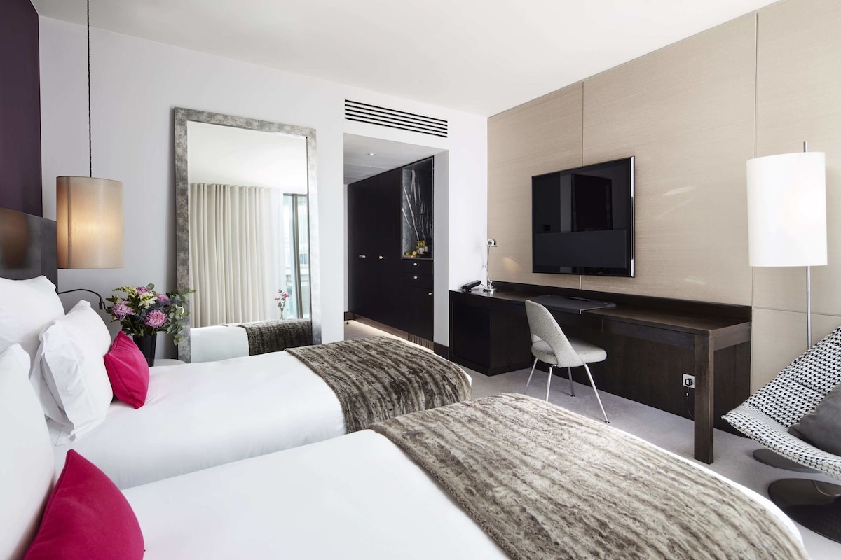 A modern hotel room with a bed, desk, chair, TV, and a large mirror.