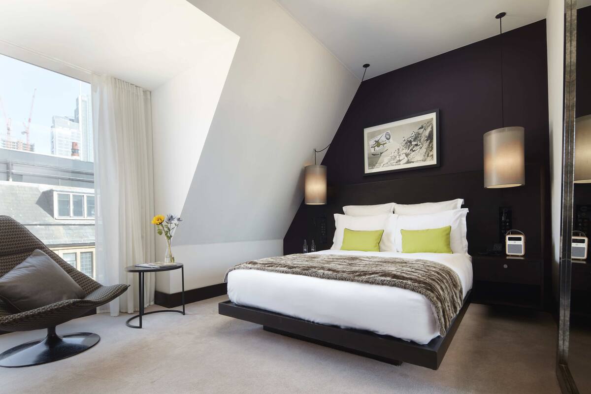 A modern hotel room with a bed, a chair, a small table with flowers, and a city view.