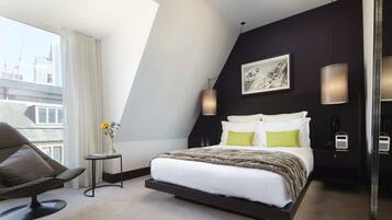 A modern hotel room with a bed, a chair, a small table with flowers, and a city view.