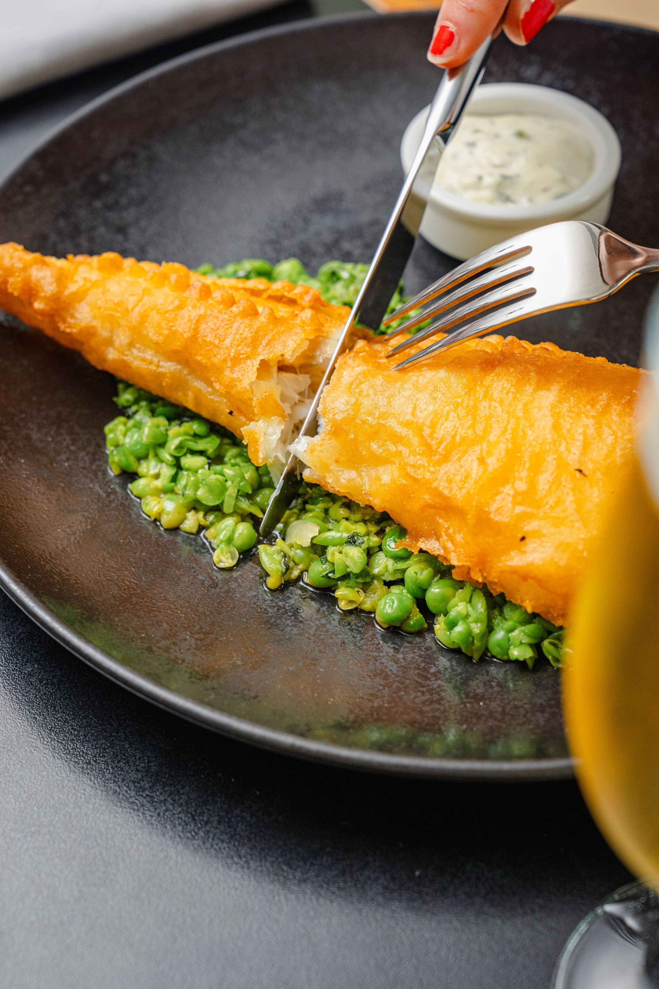Un plato con un trozo de pescado, guisantes y un recipiente pequeño de salsa.