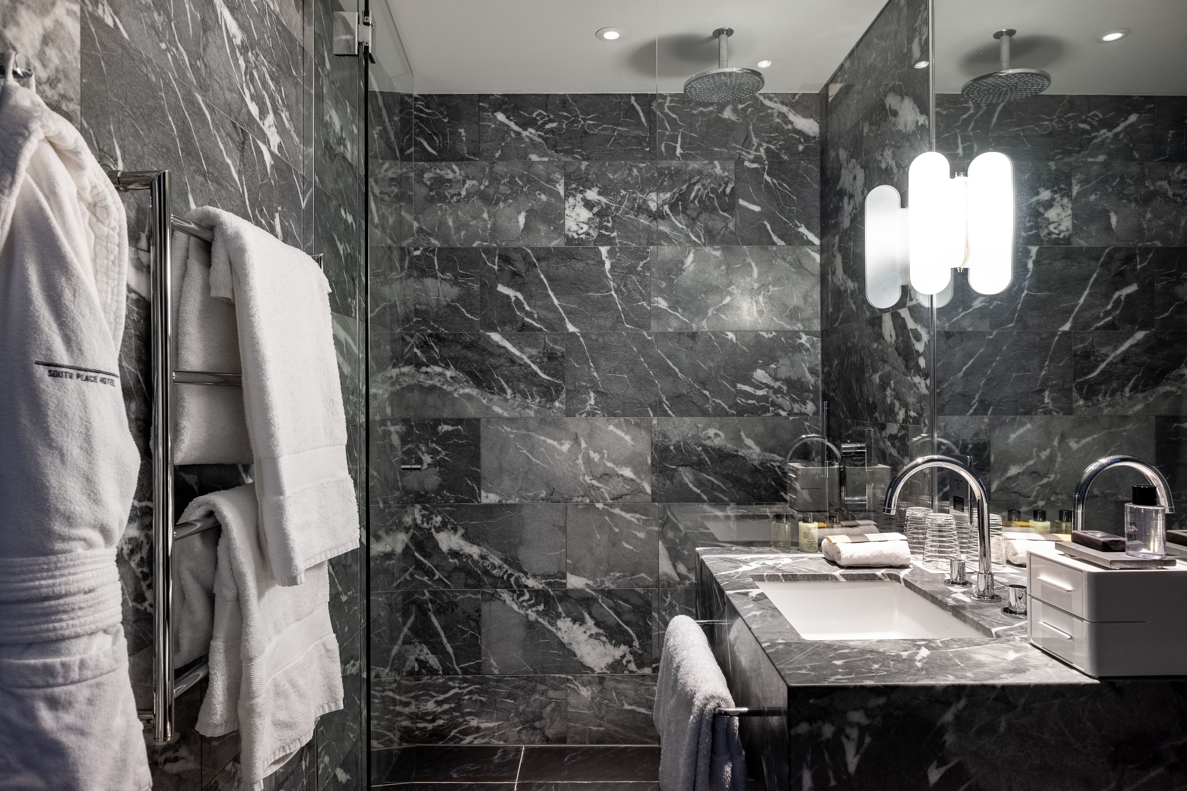 A modern bathroom with a marble wall, a glass shower enclosure, a large mirror, and a sink with a faucet.