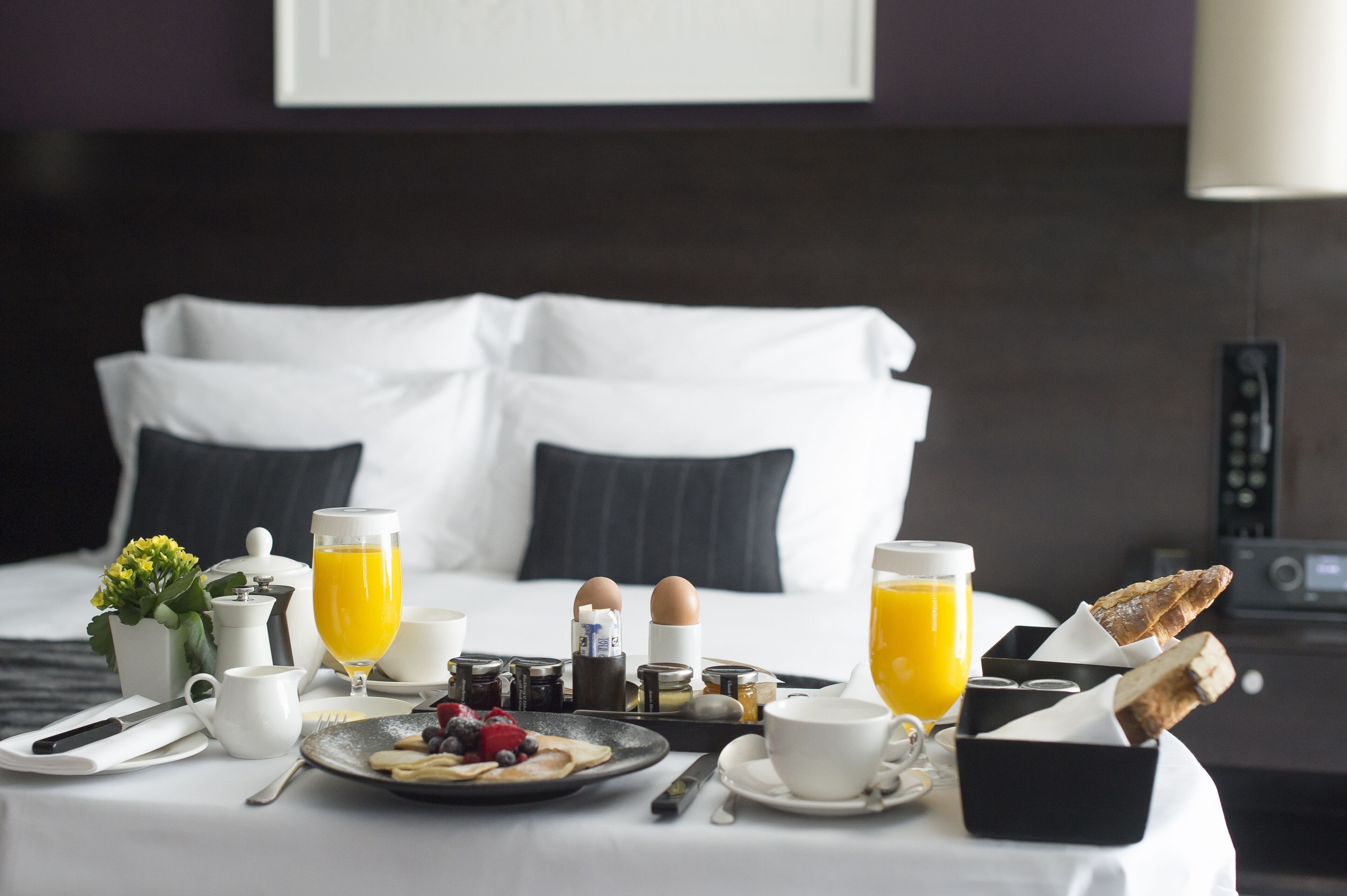 Een netjes gedekte ontbijttafel met diverse soorten eten, een koffiepot en twee glazen sinaasappelsap op een bed met witte lakens en donkere kussens.