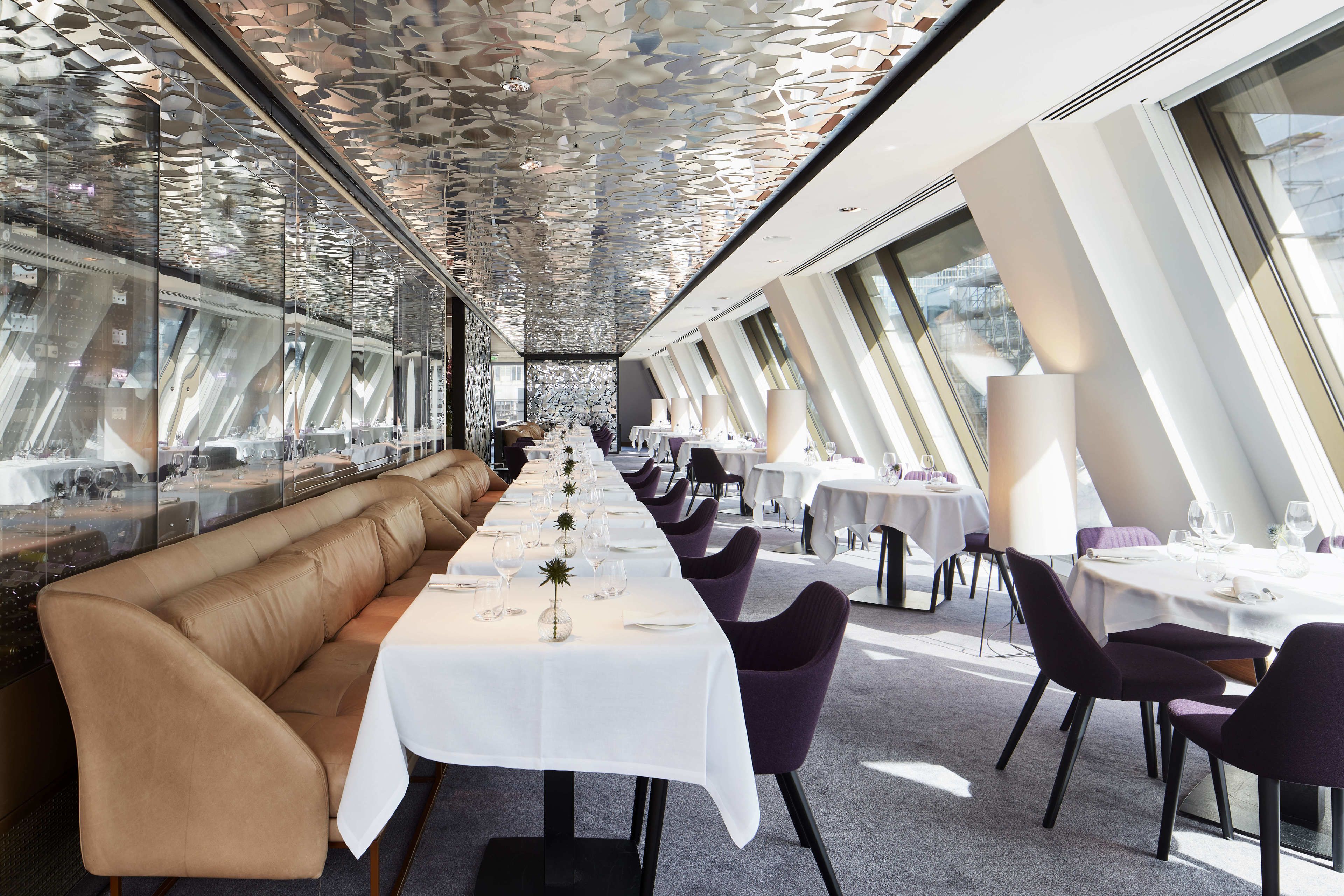 A modern restaurant with a long table, cushioned seating, and large windows.