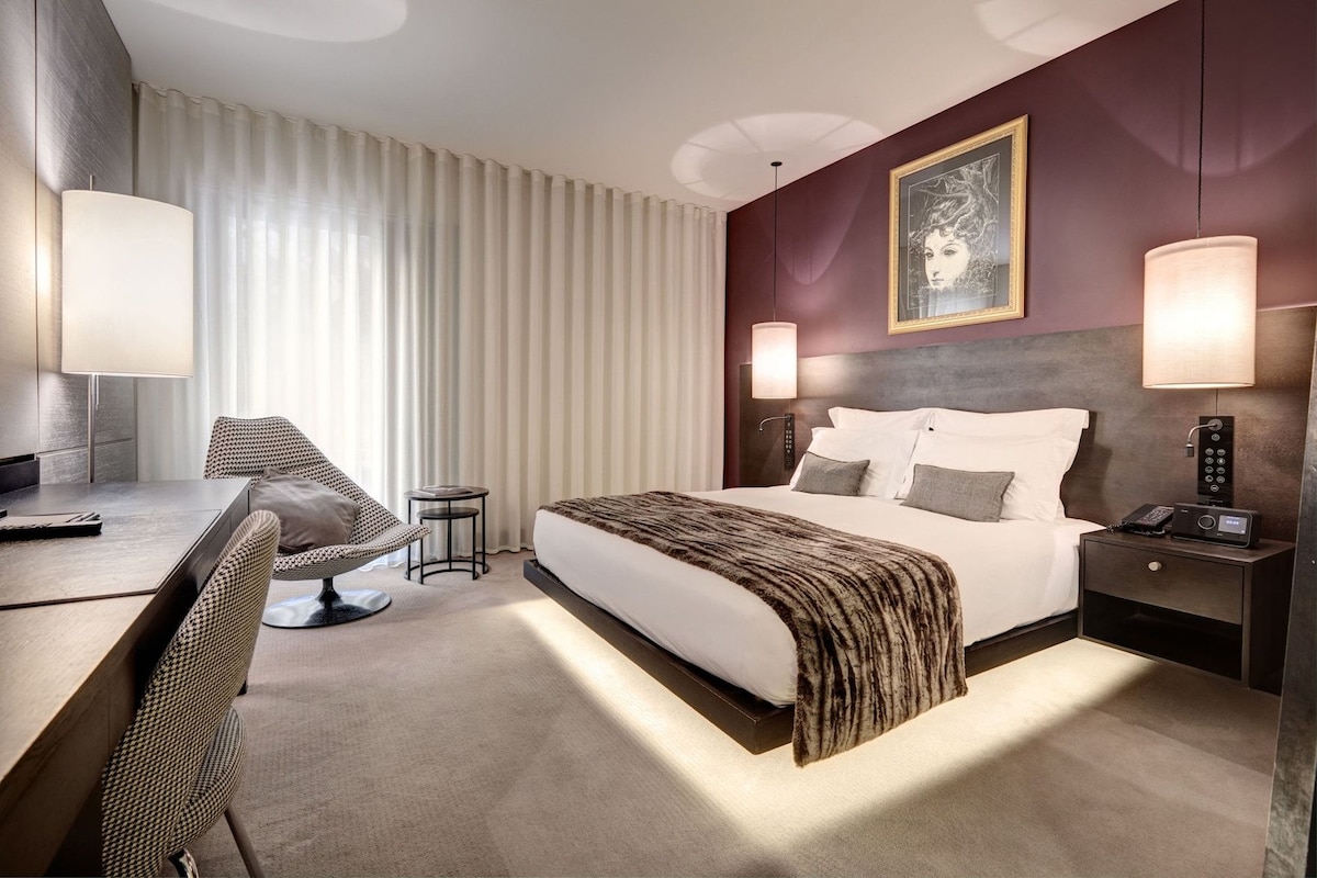 A hotel room with a bed, bedside tables, a desk, a chair, and a large window with curtains.
