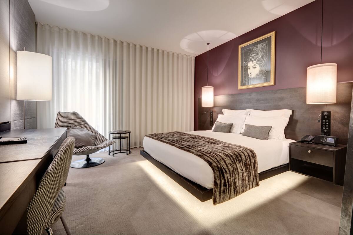 A hotel room with a large bed, a desk with a chair, a bedside table with a lamp, and a painting on the wall.