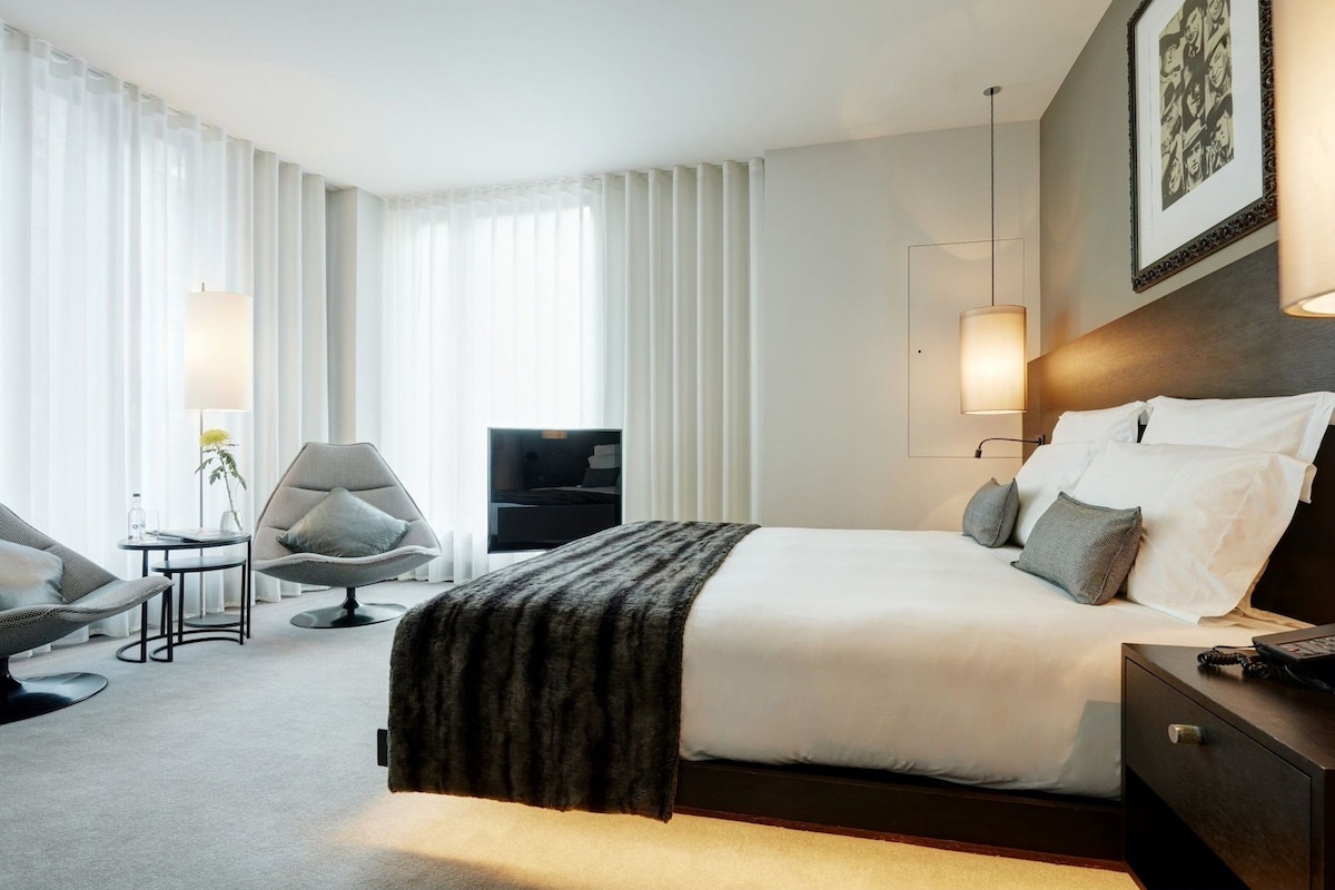 A hotel room with a large bed, a desk, a chair, a TV, and a wall-mounted lamp.