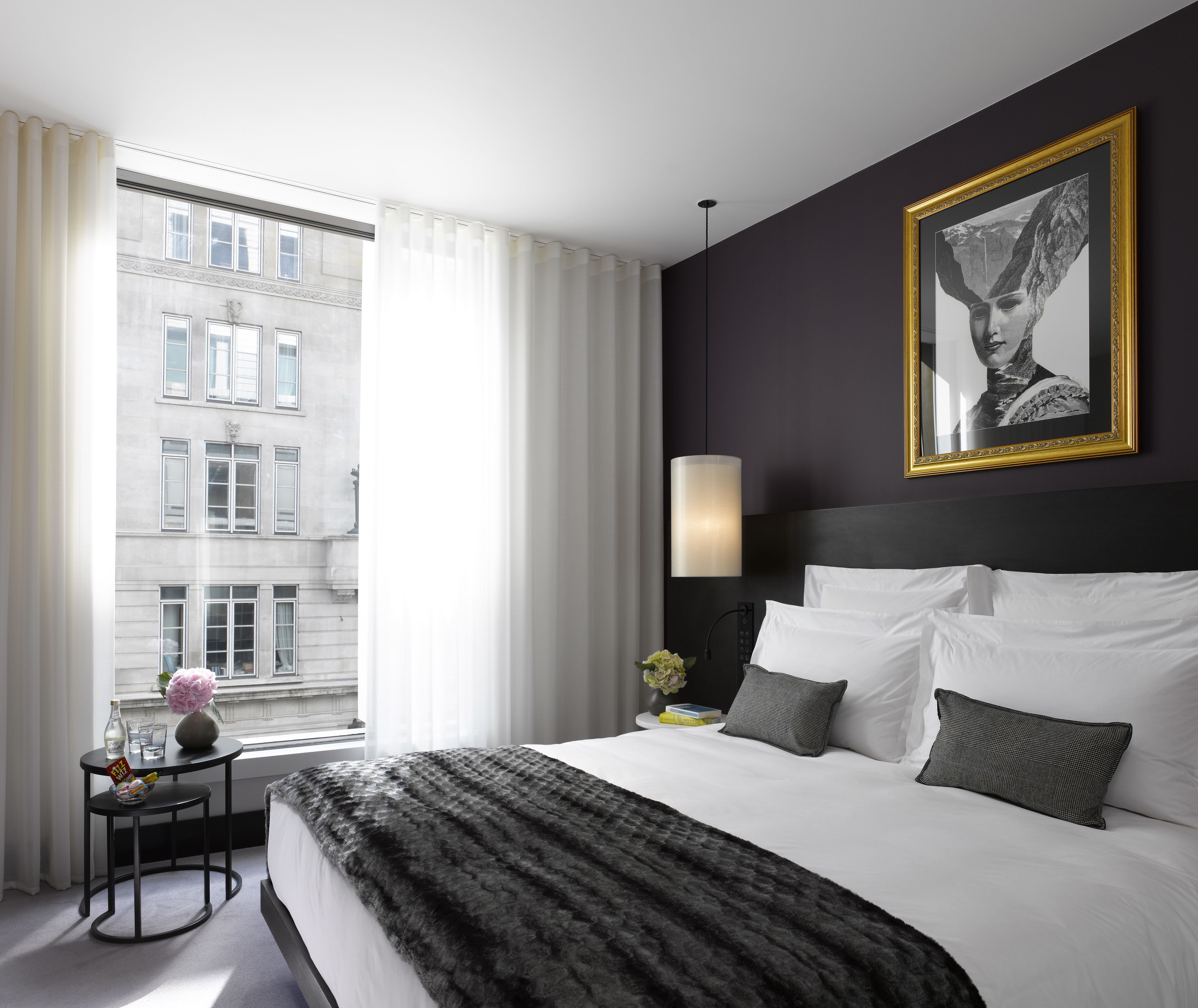 A hotel room with a large bed, a nightstand, a lamp, a framed picture on the wall, and a view of a building through the window.