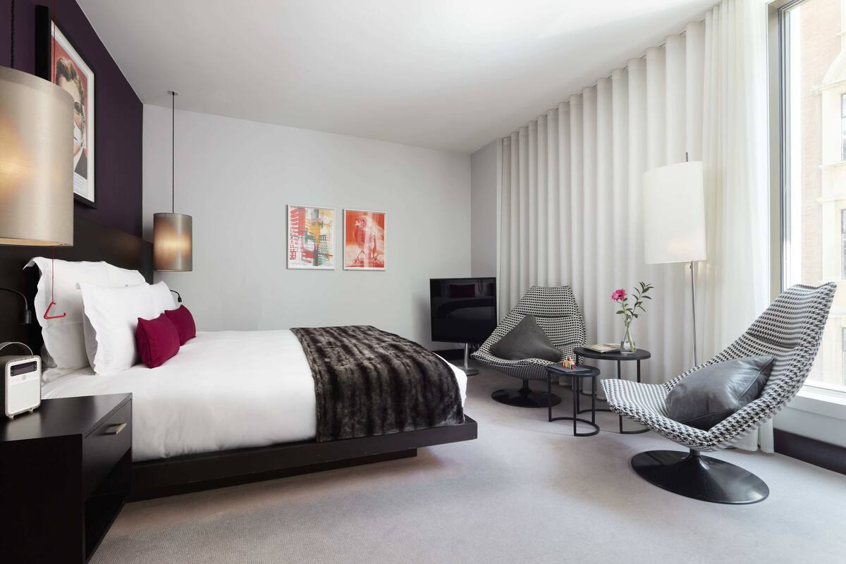 A modern hotel room with a bed, a desk, a chair, a TV, and a window with curtains.