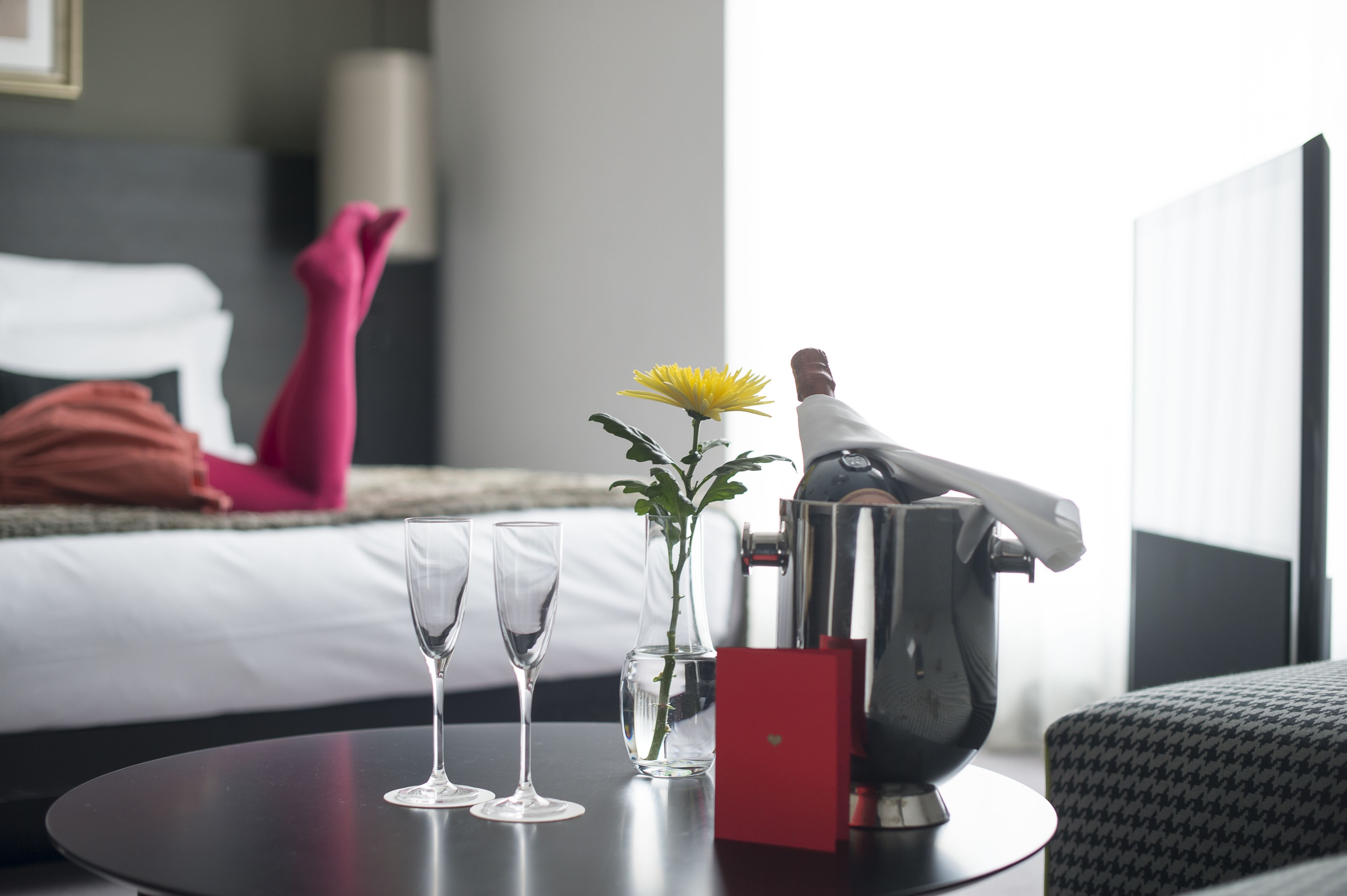 Una camera d'albergo con un letto, un comodino con un vaso di fiori, due bicchieri e un minibar.