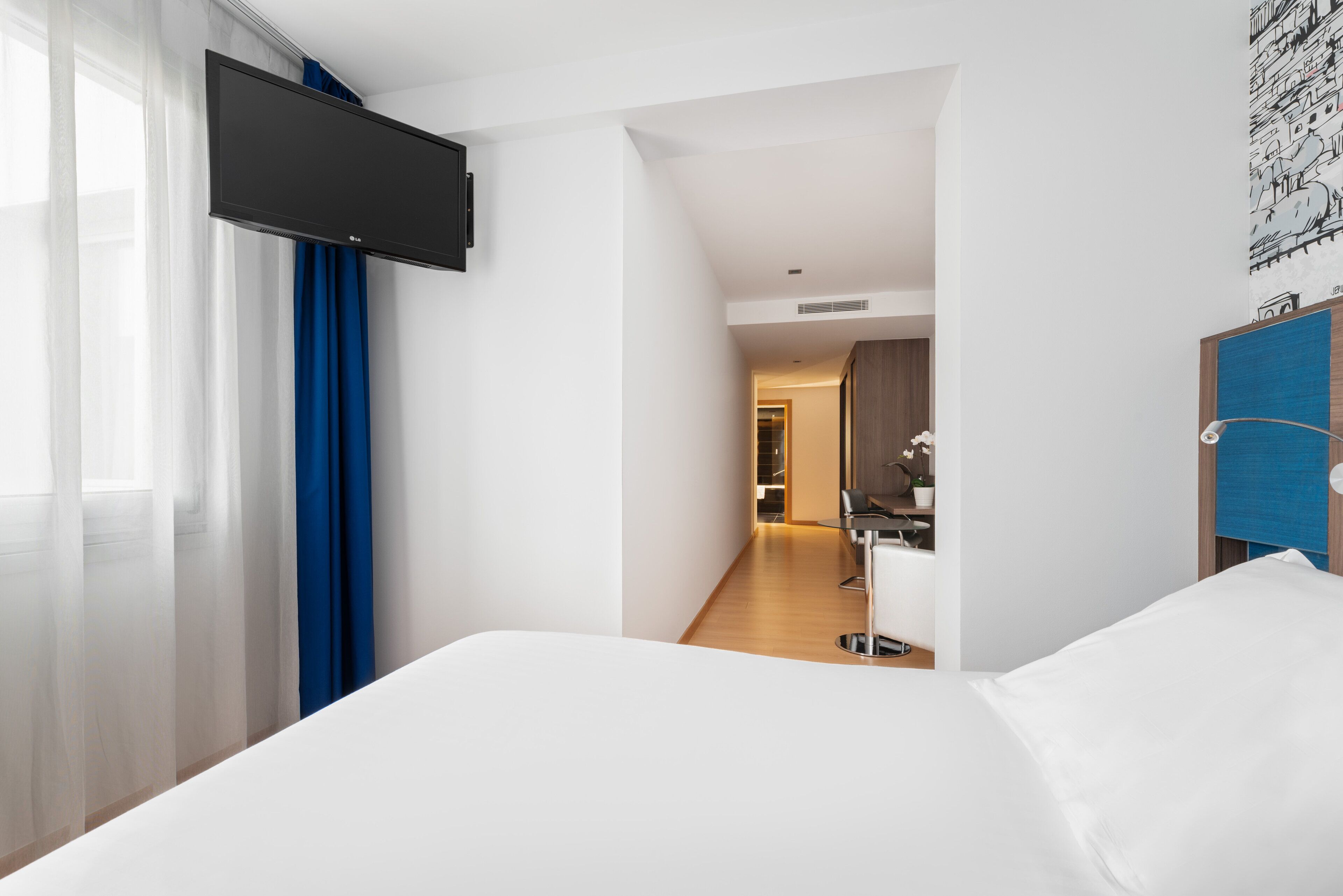 Una camera d'albergo con un letto, una TV a parete, un bagno con specchio e un corridoio che porta a un'altra stanza.