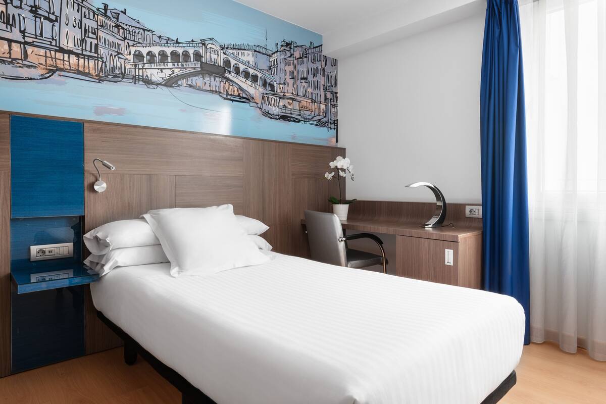 Una habitación de hotel con cama, escritorio, silla, mesita de noche, lámpara de pared y un cuadro de un paisaje urbano.