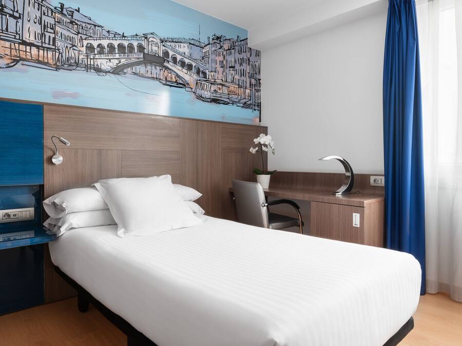 Una habitación de hotel con cama, escritorio, silla, mesita de noche, lámpara de pared y un cuadro de un paisaje urbano.
