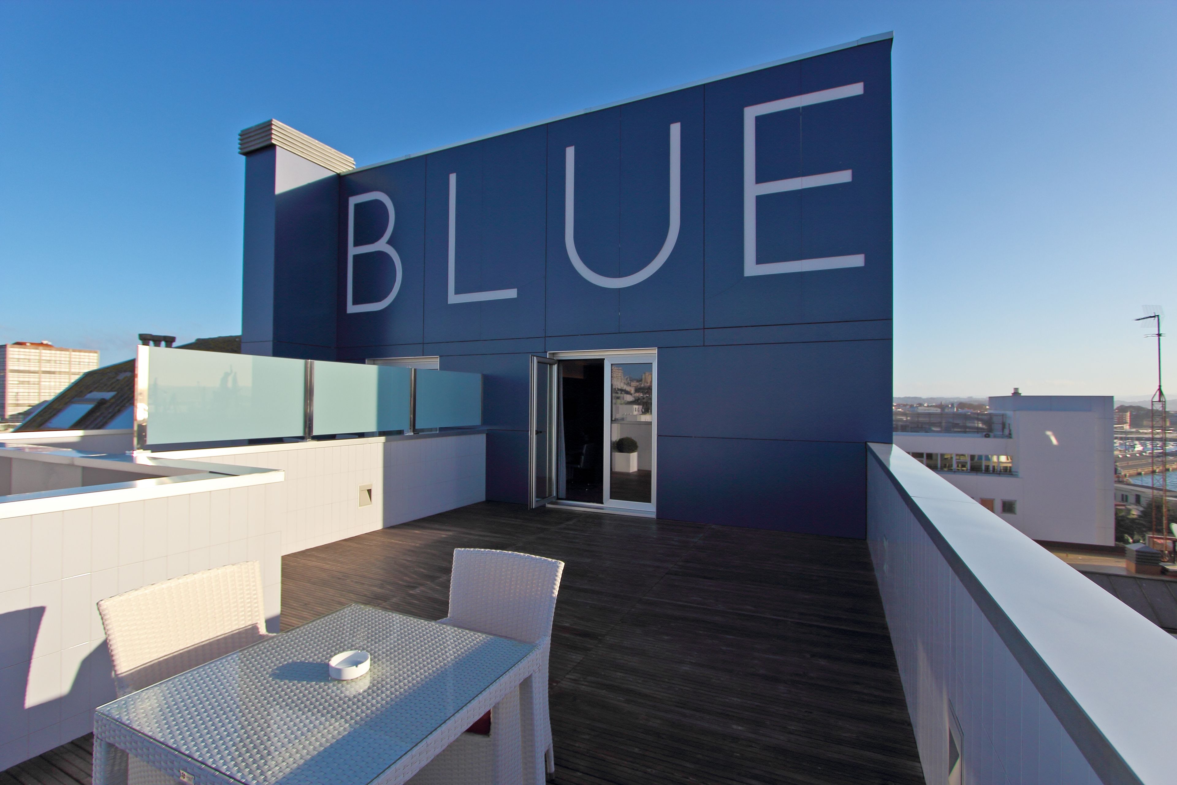 A rooftop terrace with a large 'BLUE' sign, a glass balustrade, and a table with chairs.