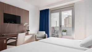 Une chambre d’hôtel comprenant un lit, un bureau avec une télévision, une chaise, un balcon avec vue sur les bâtiments et une plante en pot.