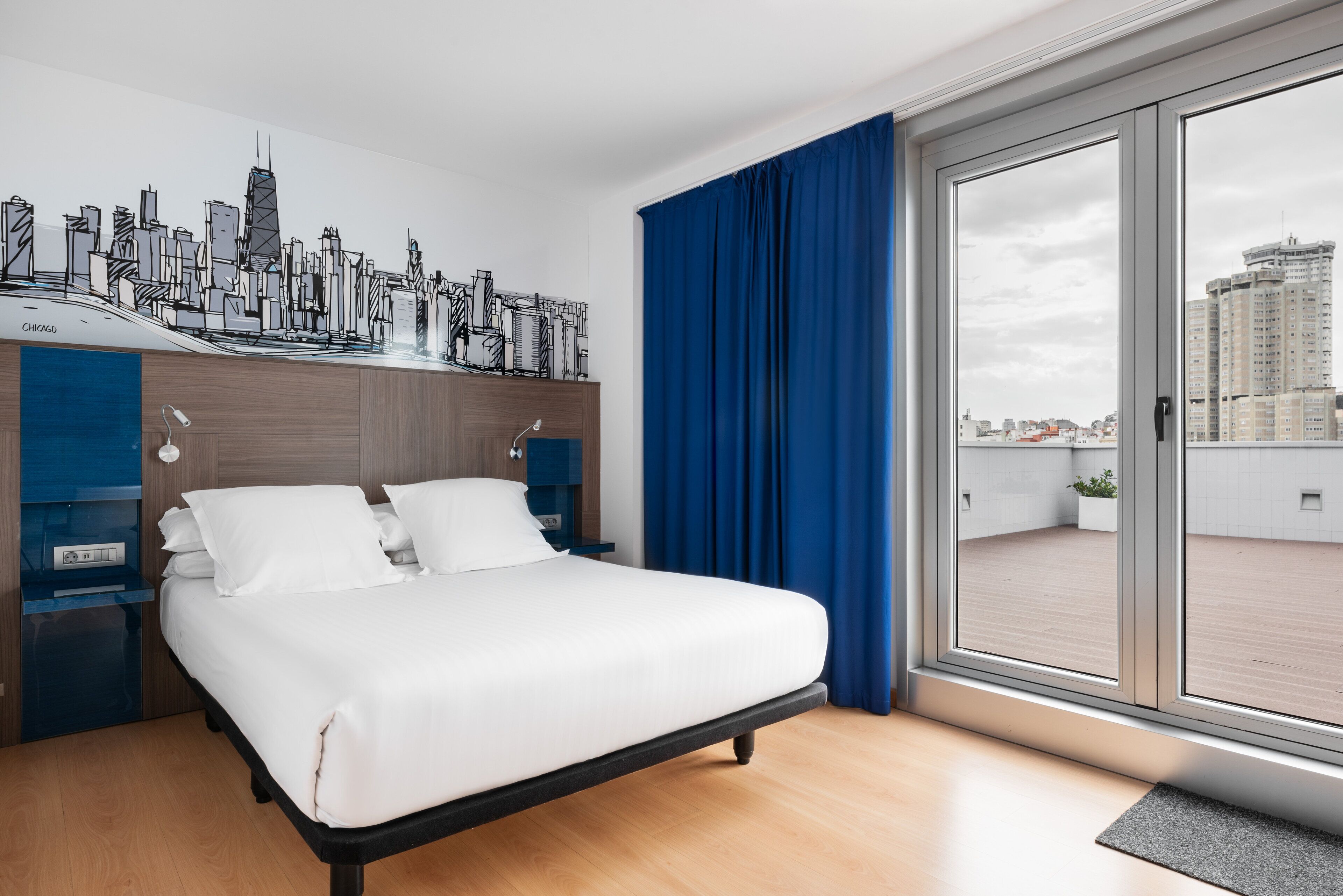 A modern hotel room with a cityscape mural, a large bed, a desk, and a sliding glass door to an outdoor balcony.