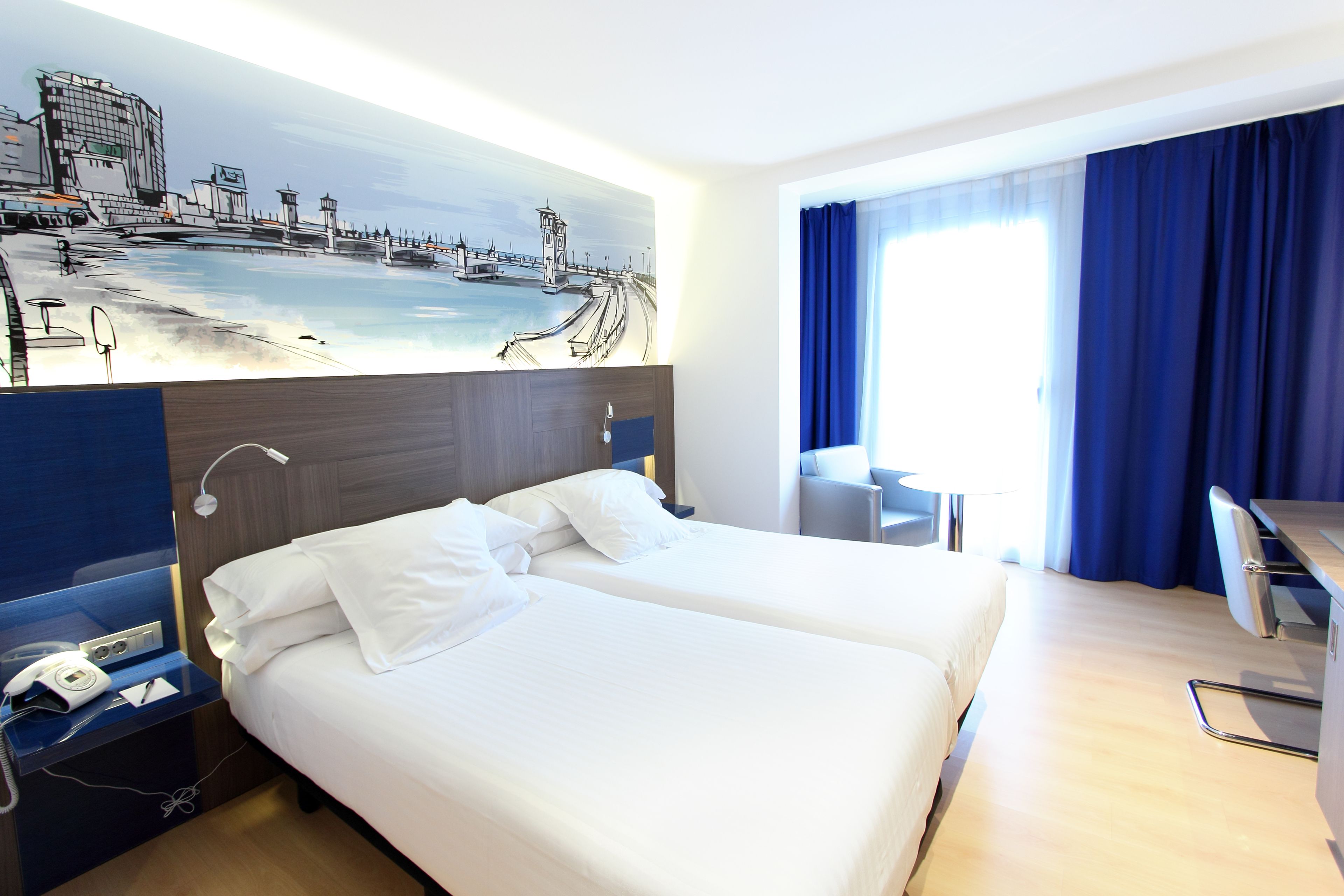 A hotel room with two beds, a desk, a chair, a painting on the wall, and a window with curtains.