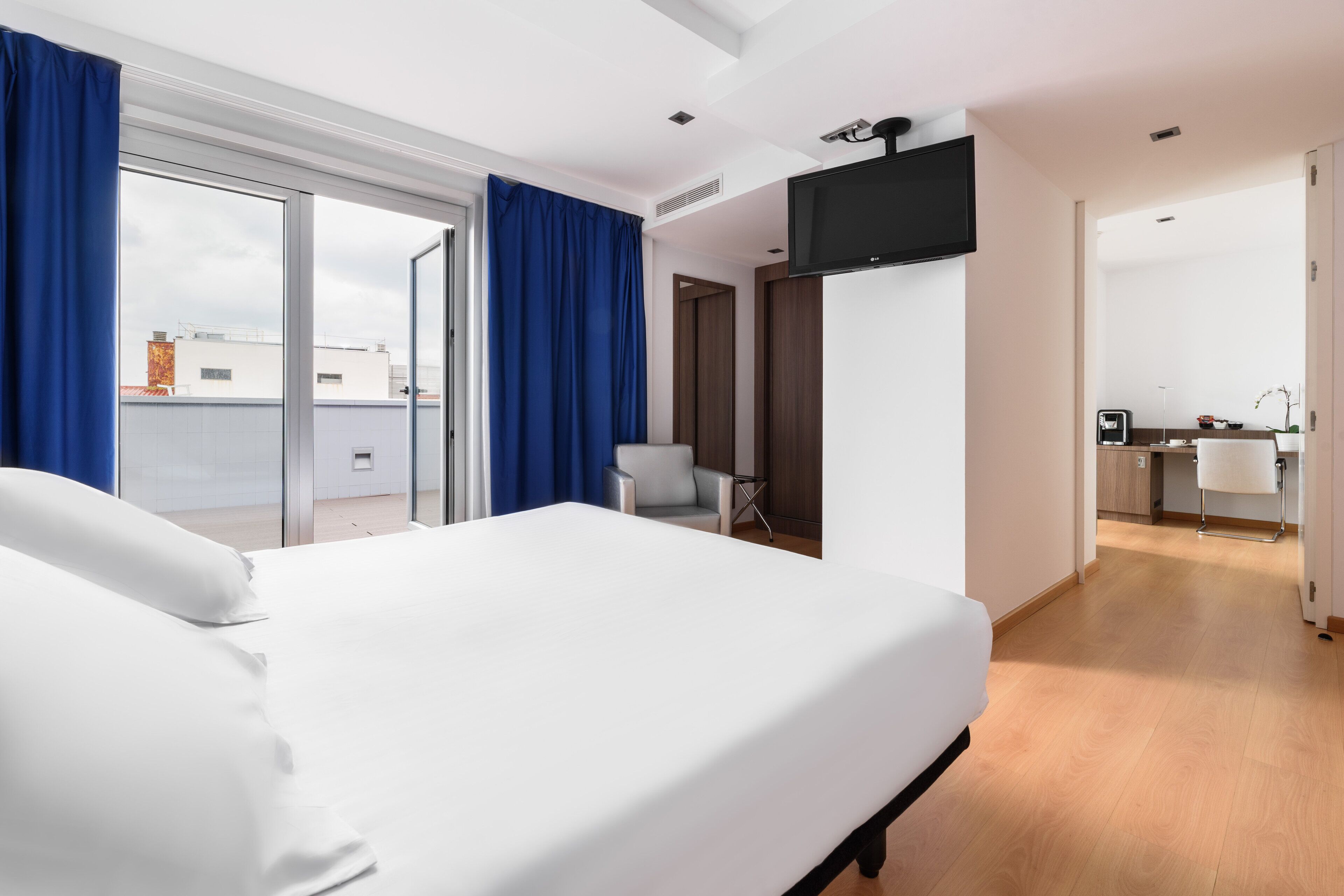 Une chambre d’hôtel moderne dotée d’un grand lit, d’un téléviseur à écran plat fixé au mur et d’une porte-fenêtre coulissante donnant sur un balcon.