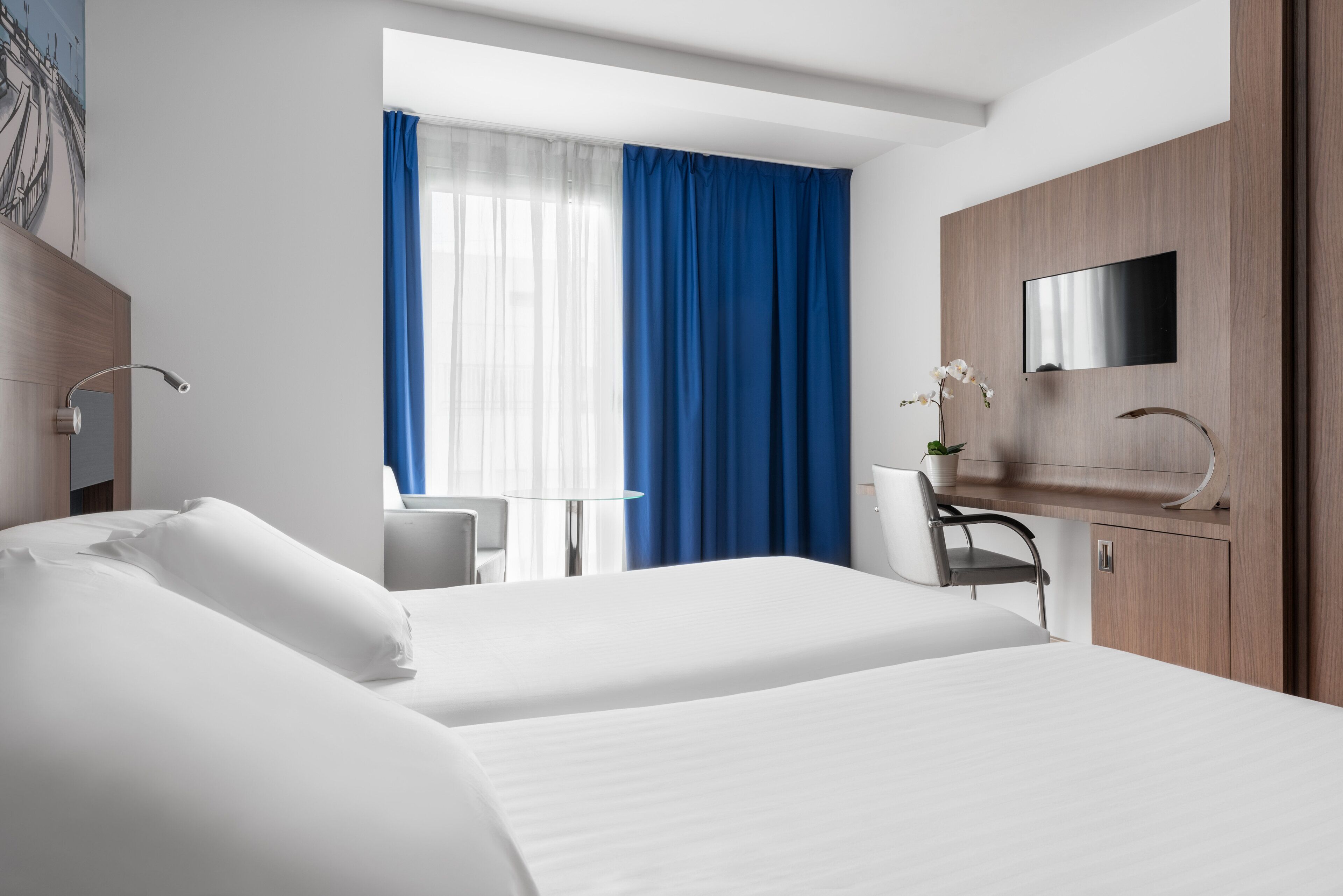 A hotel room with a bed, a desk, a chair, a TV, and a window with blue curtains.