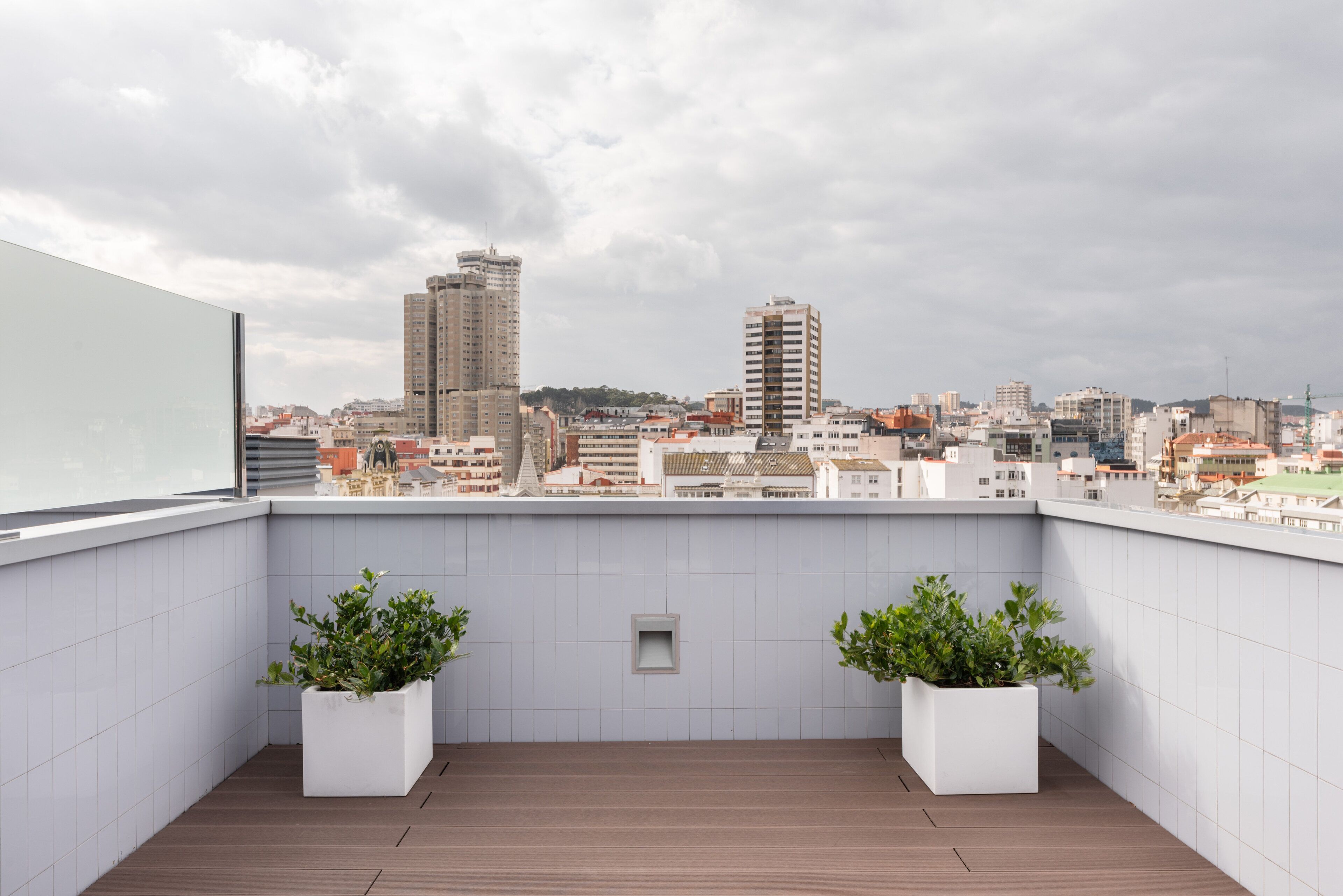 A rooftop terrace with two potted plants, a tiled wall, and a cityscape view.