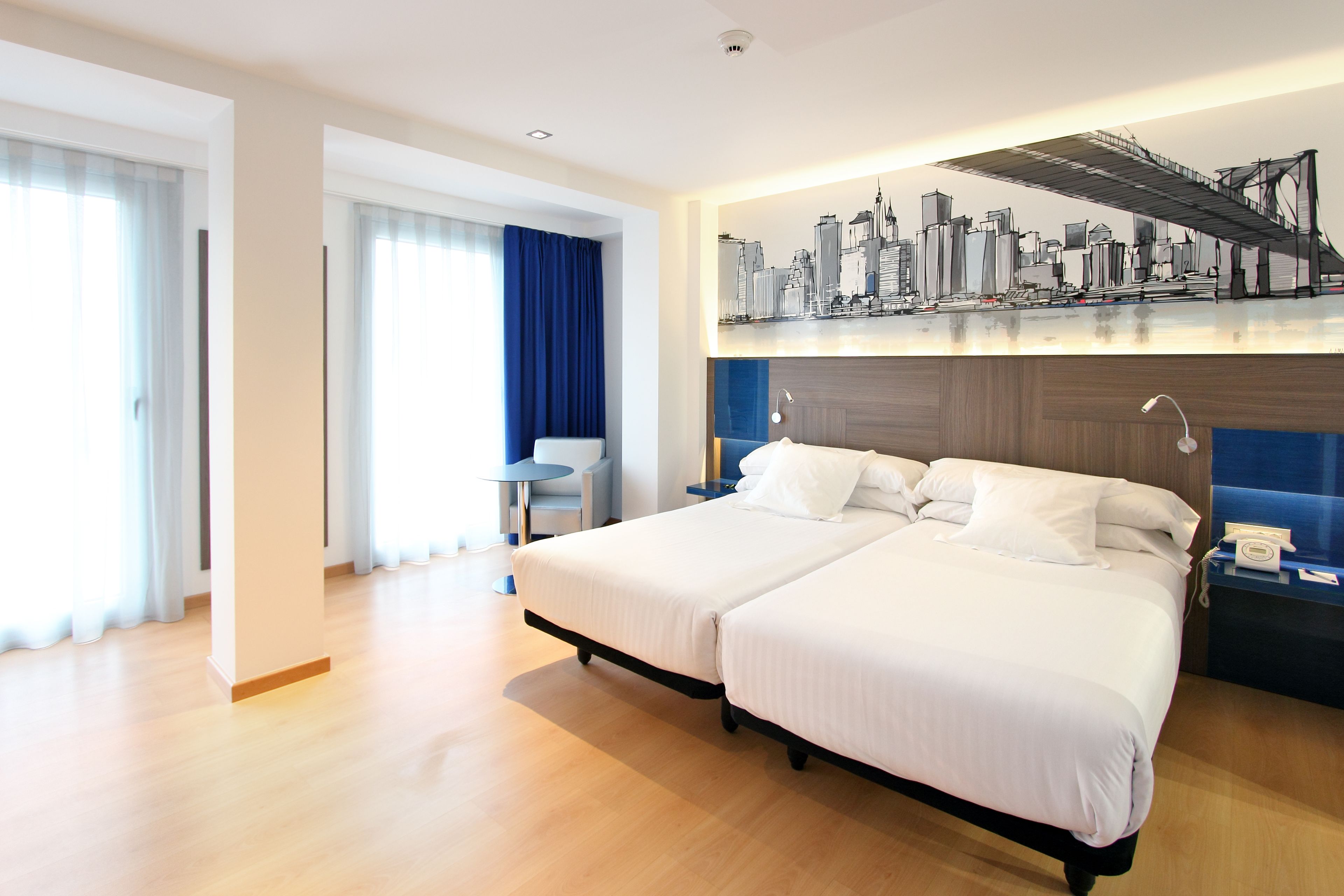 Habitación de hotel con dos camas, un mural de paisaje urbano, un escritorio y una silla.