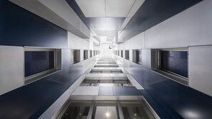 Un couloir moderne et épuré, doté d’un escalier central, flanqué de panneaux de verre et de surfaces métalliques.