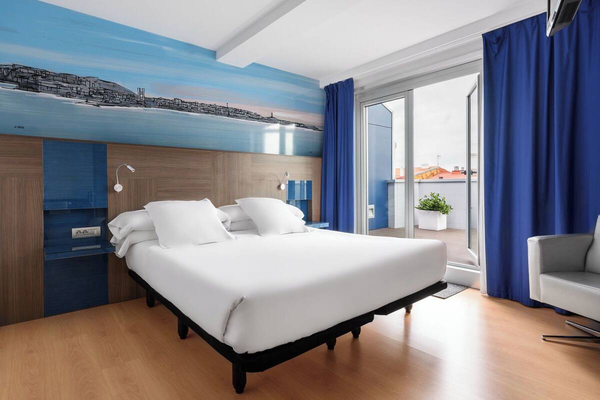 Habitación de hotel moderna con una cama, un escritorio, una silla y vistas de la ciudad a través de un amplio ventanal.