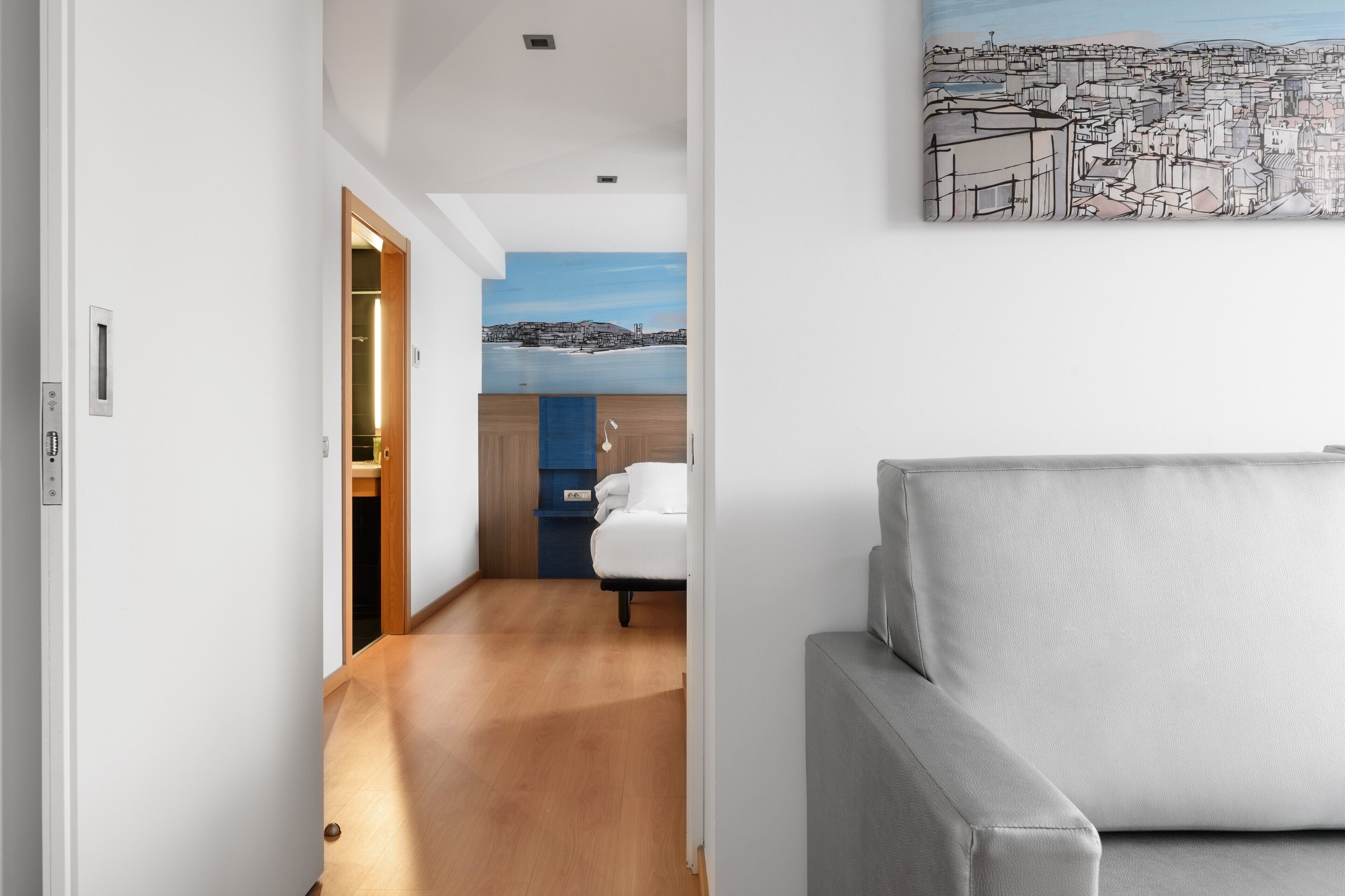Moderni hotellihuone, jossa on puulattia, valkoinen sohva, sininen kaappi ja kaupunkimaisemaa esittävä taulu seinällä.
