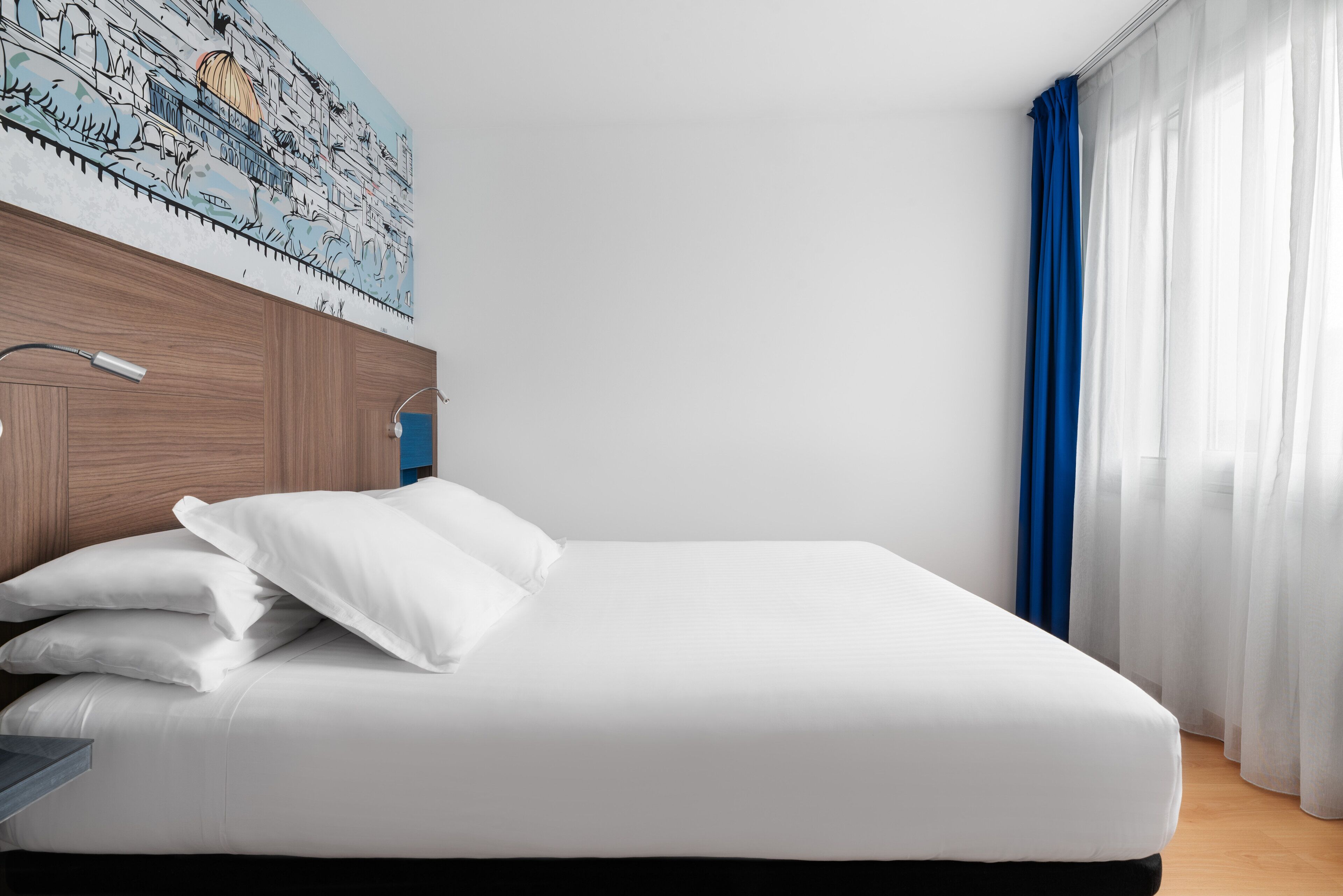 A neatly made bed with white linens and pillows, a wooden headboard, and a wall-mounted lamp.