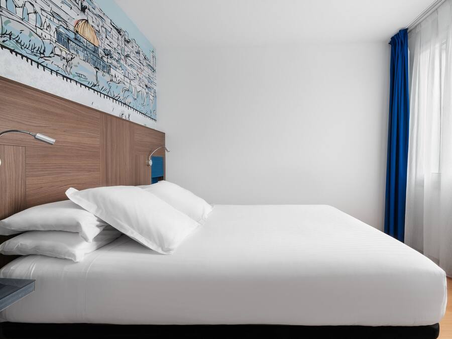 Una cama bien hecha con sábanas y almohadas blancas, un cabecero de madera y una lámpara de pared.