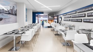 Un espace de restauration moderne avec des tables et des chaises blanches, une fresque représentant un paysage urbain et des tableaux encadrés accrochés aux murs.