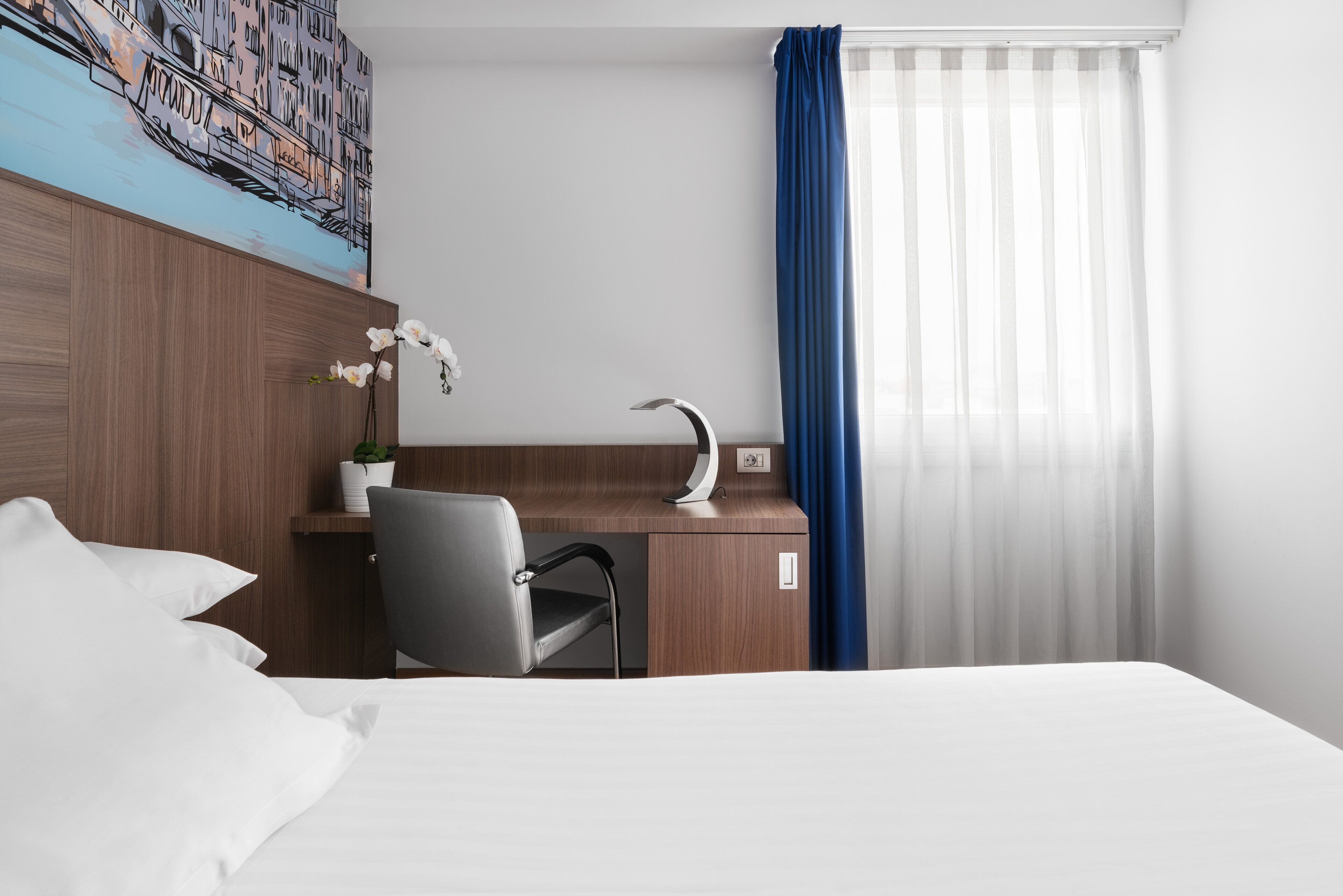 A hotel room with a bed, a desk with a chair, a potted plant, a painting on the wall, and a window with curtains.
