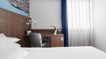 Habitación de hotel con cama, escritorio con silla, planta en maceta, un cuadro en la pared y ventana con cortinas.