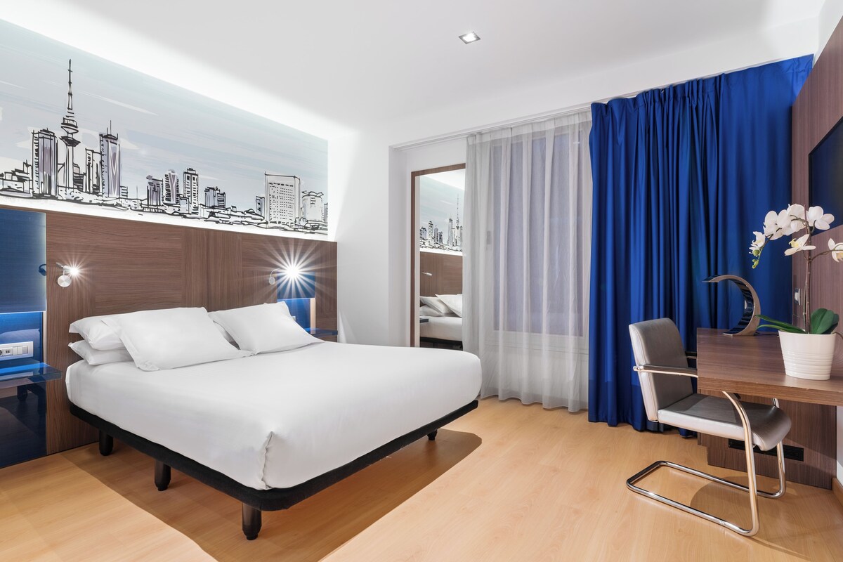 Une chambre d’hôtel moderne équipée d’un lit, d’un bureau, d’une chaise et d’une fresque murale représentant un paysage urbain.