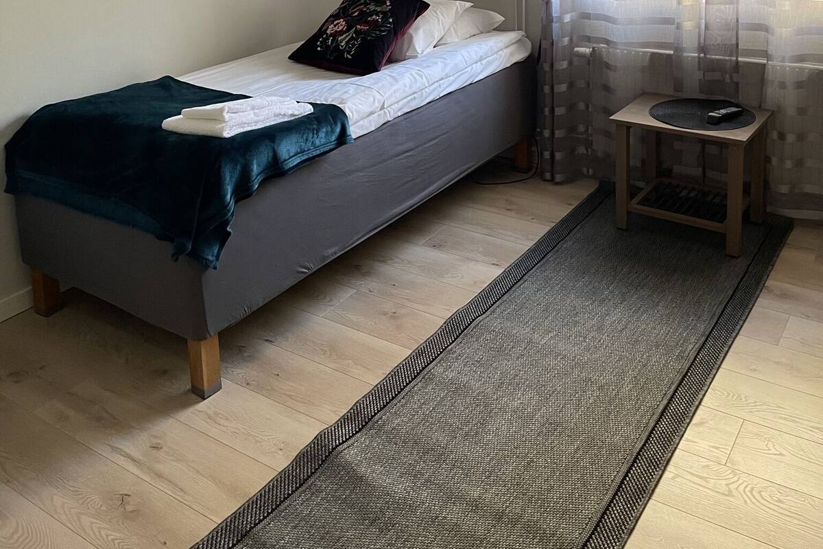 Un lit simple avec une couverture et des oreillers sombres, une petite table de chevet avec une télécommande et une fenêtre ornée de rideaux légers.