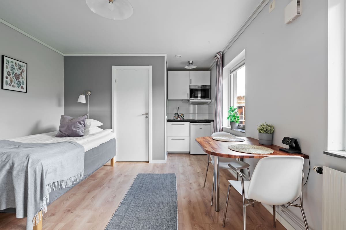 Une pièce compacte et moderne, comprenant un lit, une petite table à manger, une chaise, un coin cuisine et une fenêtre donnant sur un paysage agréable.