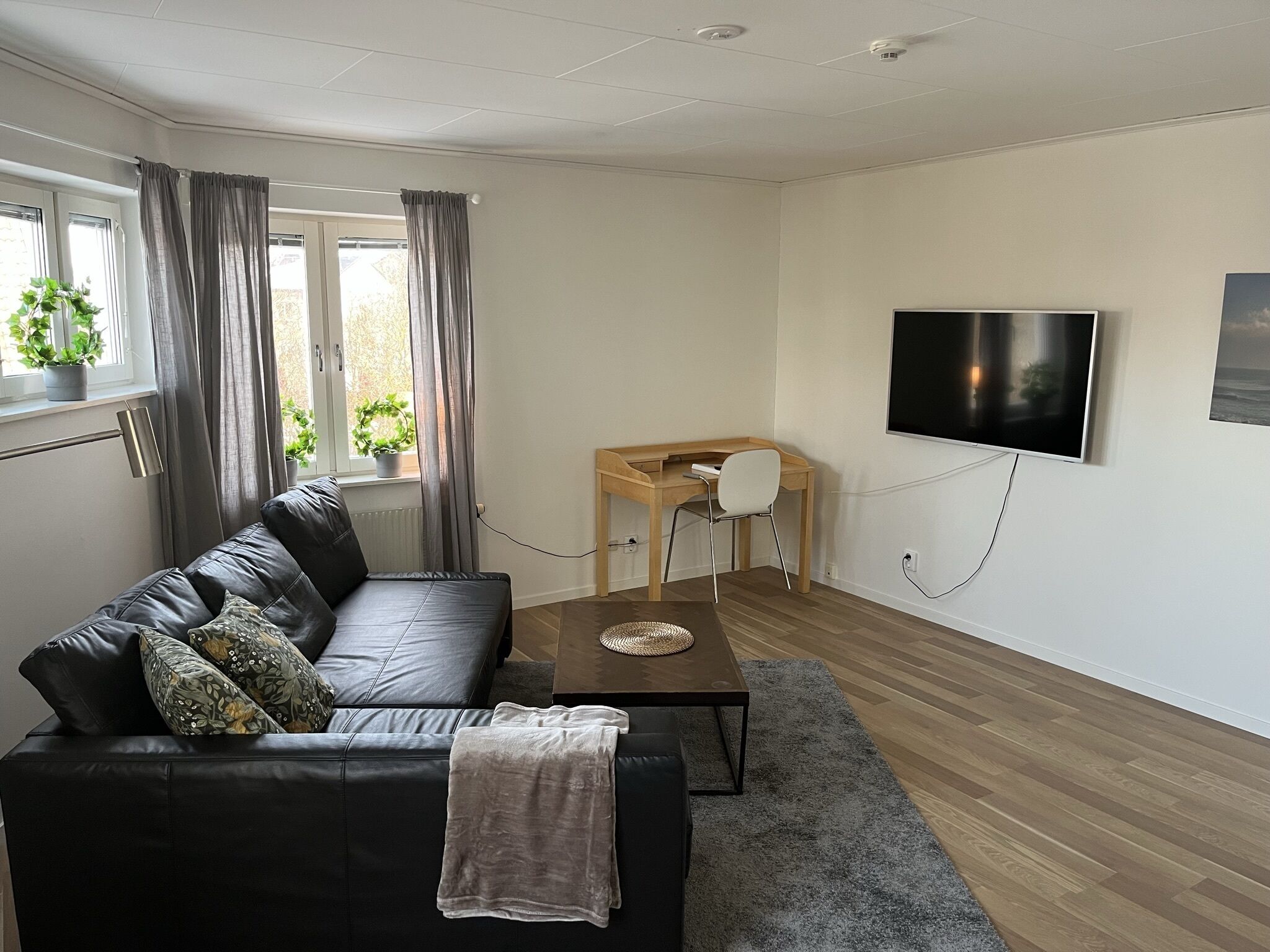 A modern living room with a black leather sofa, a wooden coffee table, a TV mounted on the wall, and a small wooden desk.