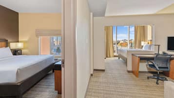 A hotel room with a bed, desk, chair, and a large window offering a city view.