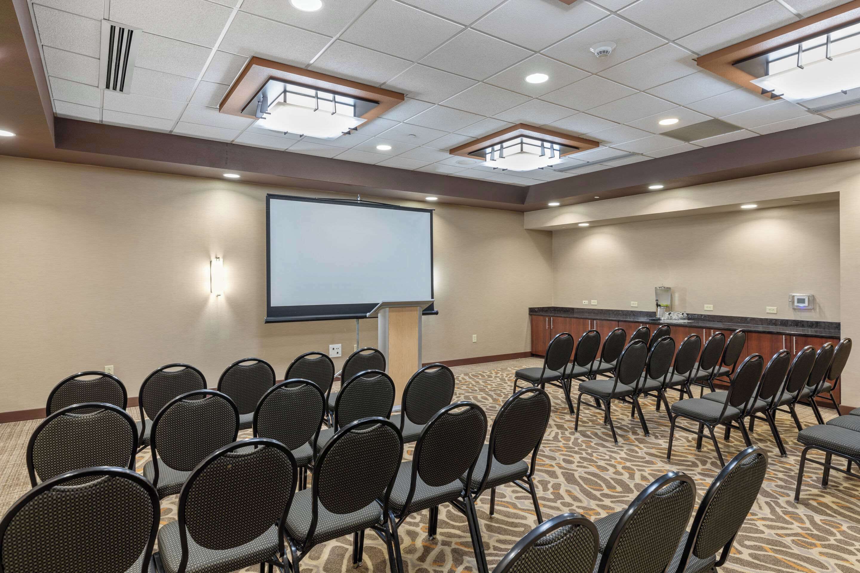 A conference room with rows of chairs facing a projector screen, a podium, and a counter with a sink.