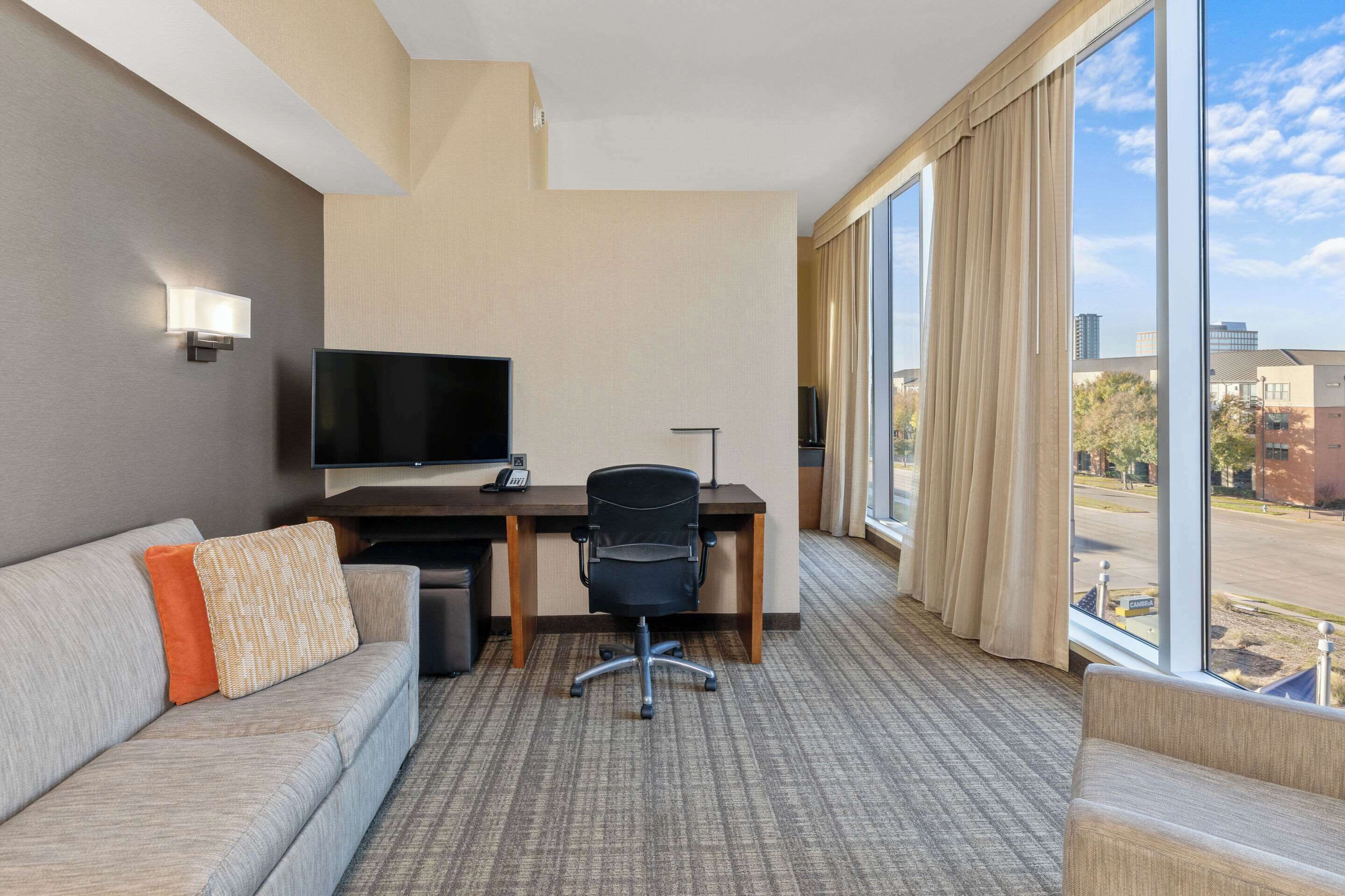 Une chambre d’hôtel dotée d’une grande fenêtre, d’un téléviseur à écran plat, d’un bureau avec une chaise et d’un canapé avec des coussins.