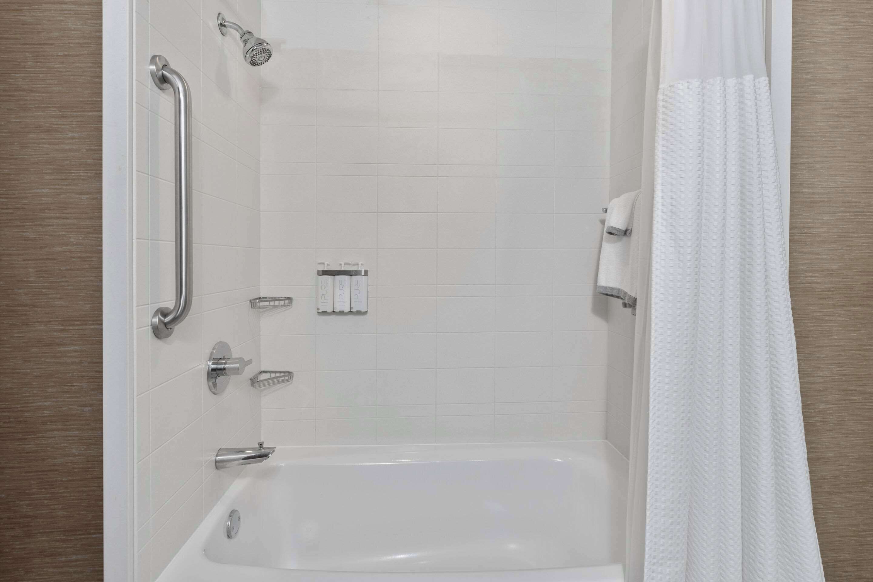 Une salle de bain moderne dotée d’une douche à l’italienne, de barres d’appui, d’une baignoire et d’un rideau de douche blanc.
